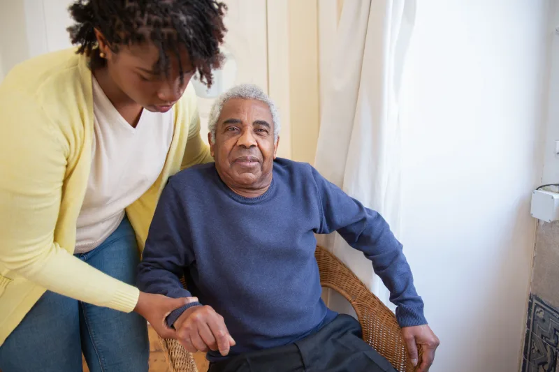 Woman gently supporting an older man seated beside a bright window