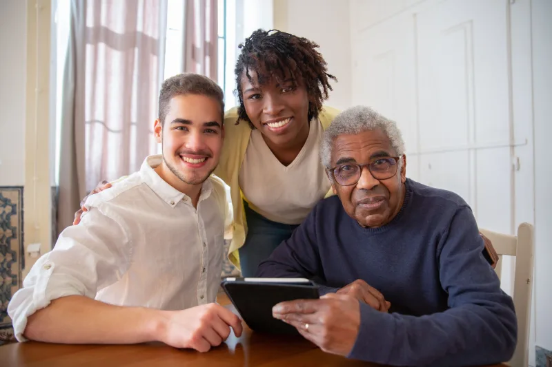 Older man and two younger adults smiling together around a tablet indoors